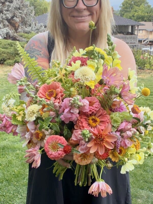 A smiling person with glasses and tattoos holds a large vibrant bouquet of mixed flowers, including pink, yellow, and white blooms, in an outdoor garden setting.