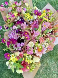 Several vibrant flower bouquets wrapped in brown paper are arranged on green grass, featuring pink, purple, yellow, and white blooms with various types of flowers and green leaves.
