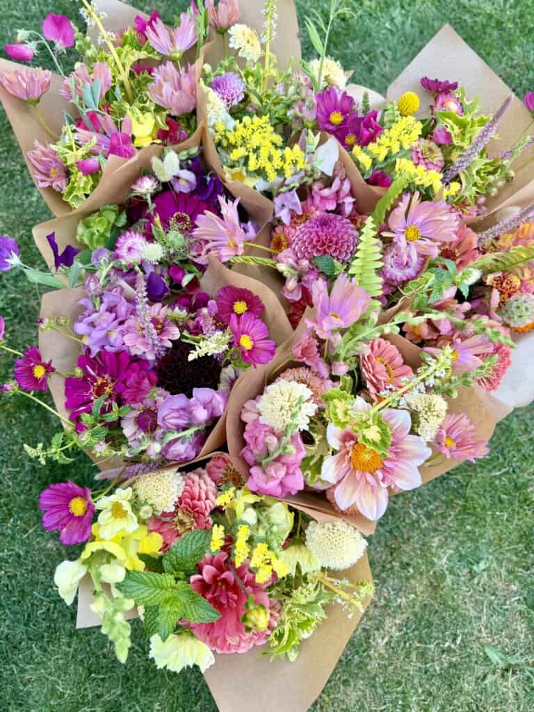 Several vibrant flower bouquets wrapped in brown paper are arranged on green grass, featuring pink, purple, yellow, and white blooms with various types of flowers and green leaves.
