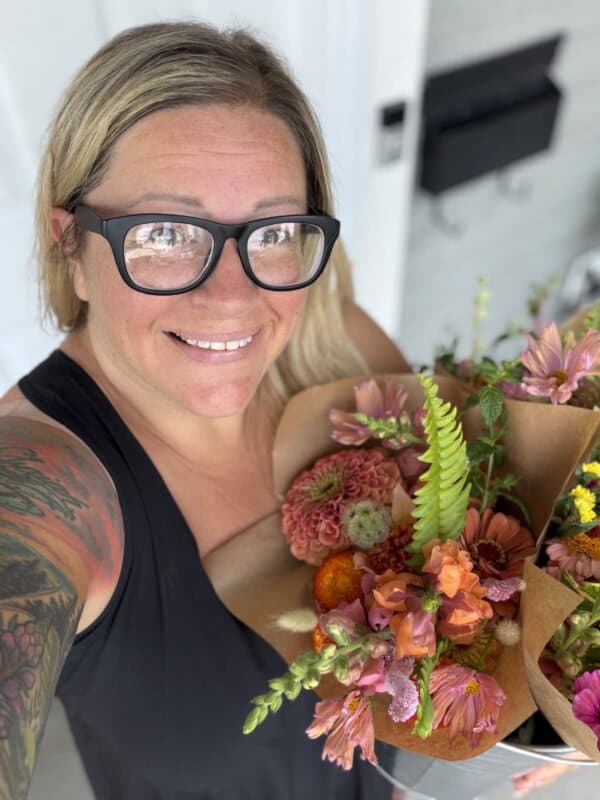 A smiling person with glasses and a tattooed arm holds colorful flower bouquets wrapped in brown paper, standing outside near a white door and mailbox.