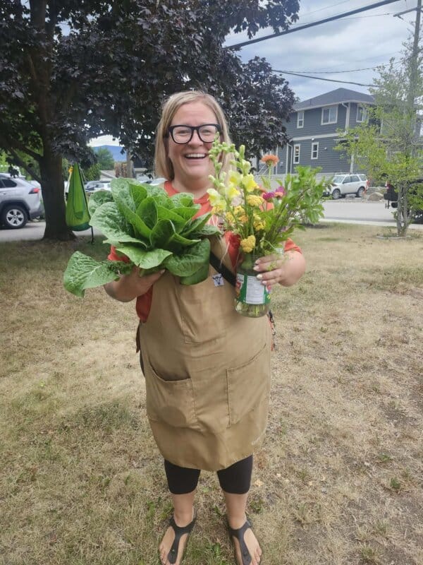 A smiling woman wearing glasses and an apron stands outside on grass, holding leafy greens in one hand and a bouquet of flowers in the other, with houses and trees in the background.